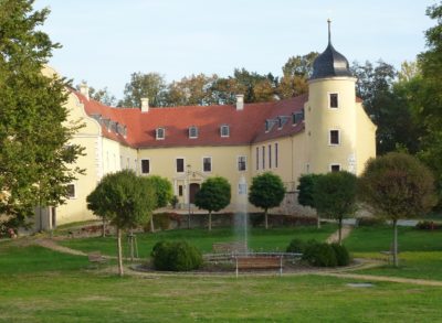 Schloss Ebersbach im Hintergrund. Im Vorderrund grüner Rasen mit rundem Springbrunnen. Hellblauer Himmel.